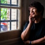 A thoughtful senior woman gazes out a window, reflecting softly indoors.